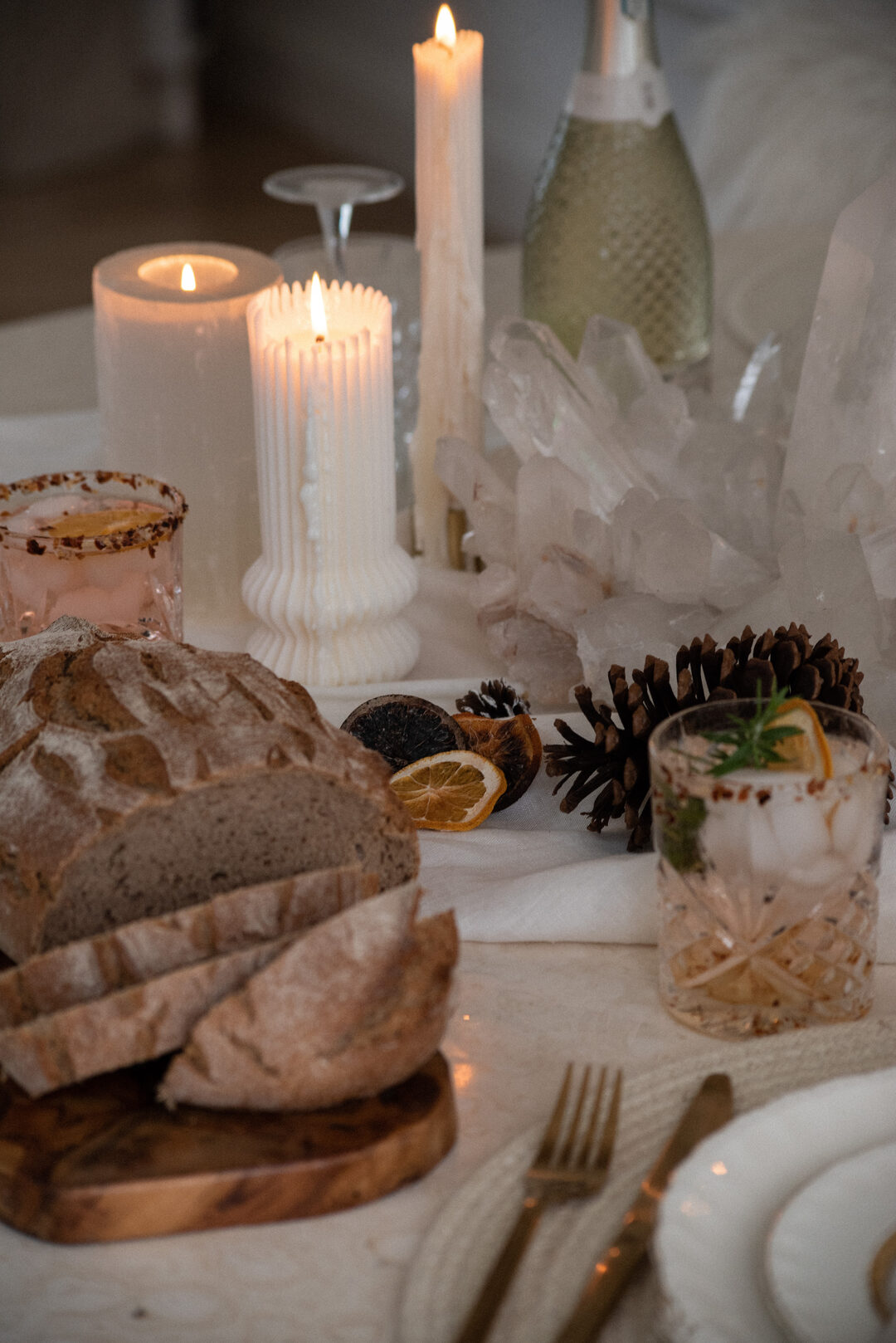 Candlelit holiday dinner table with crusty bread, sparkling wine, festive cocktails, pinecones, and dried oranges