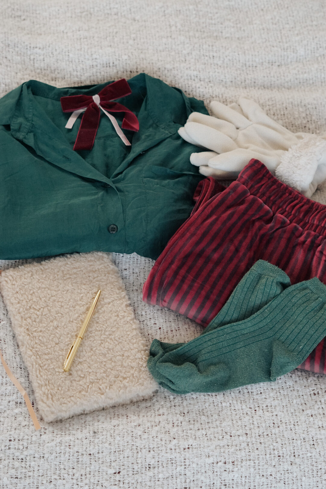 Holiday flat lay of a green silk shirt with a burgundy velvet bow, striped velvet pants, green socks, white gloves, and a sherpa notebook with a gold pen