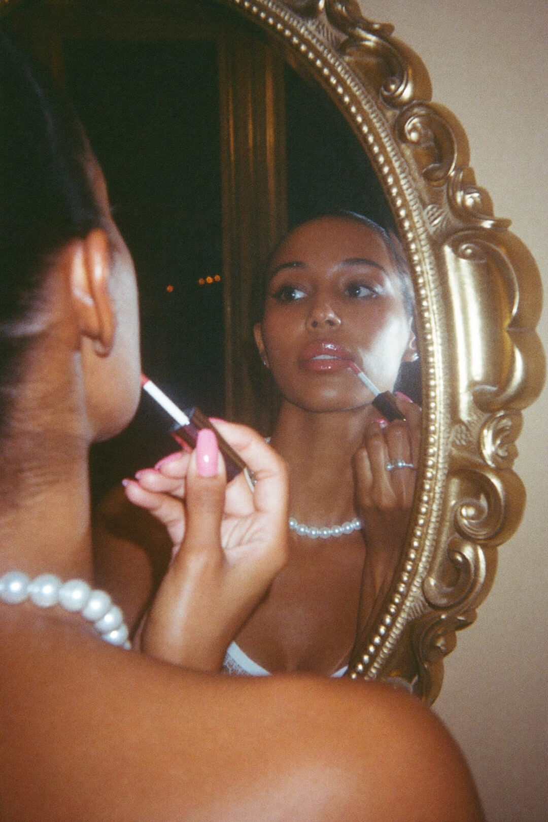 Woman with pink manicured nails applying lip gloss in an ornate gold oval mirror, wearing a pearl necklace