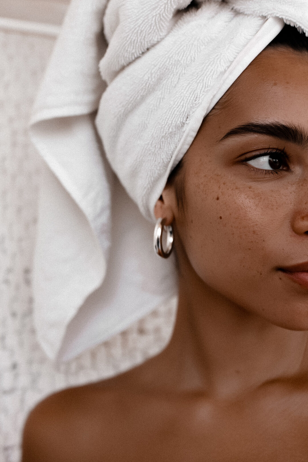 Close-up of a woman with freckled, glowing skin and a white towel wrapped around her hair, wearing silver hoop earrings