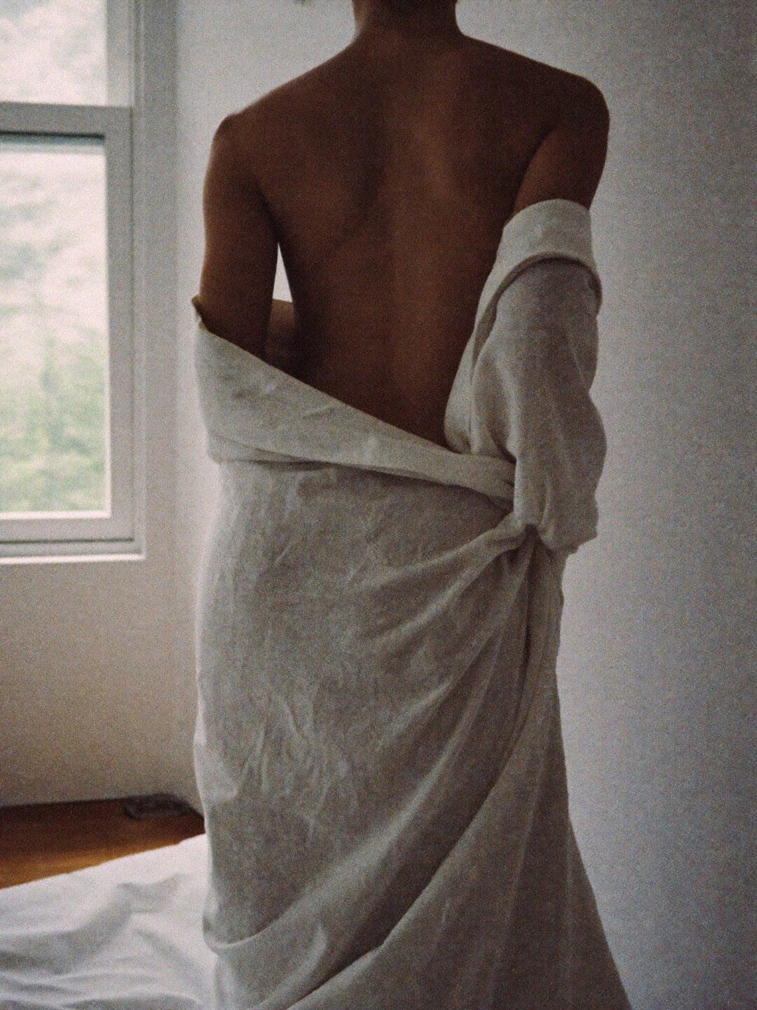 Woman with her back to the camera, wrapped in a loose linen sheet beside a softly lit window