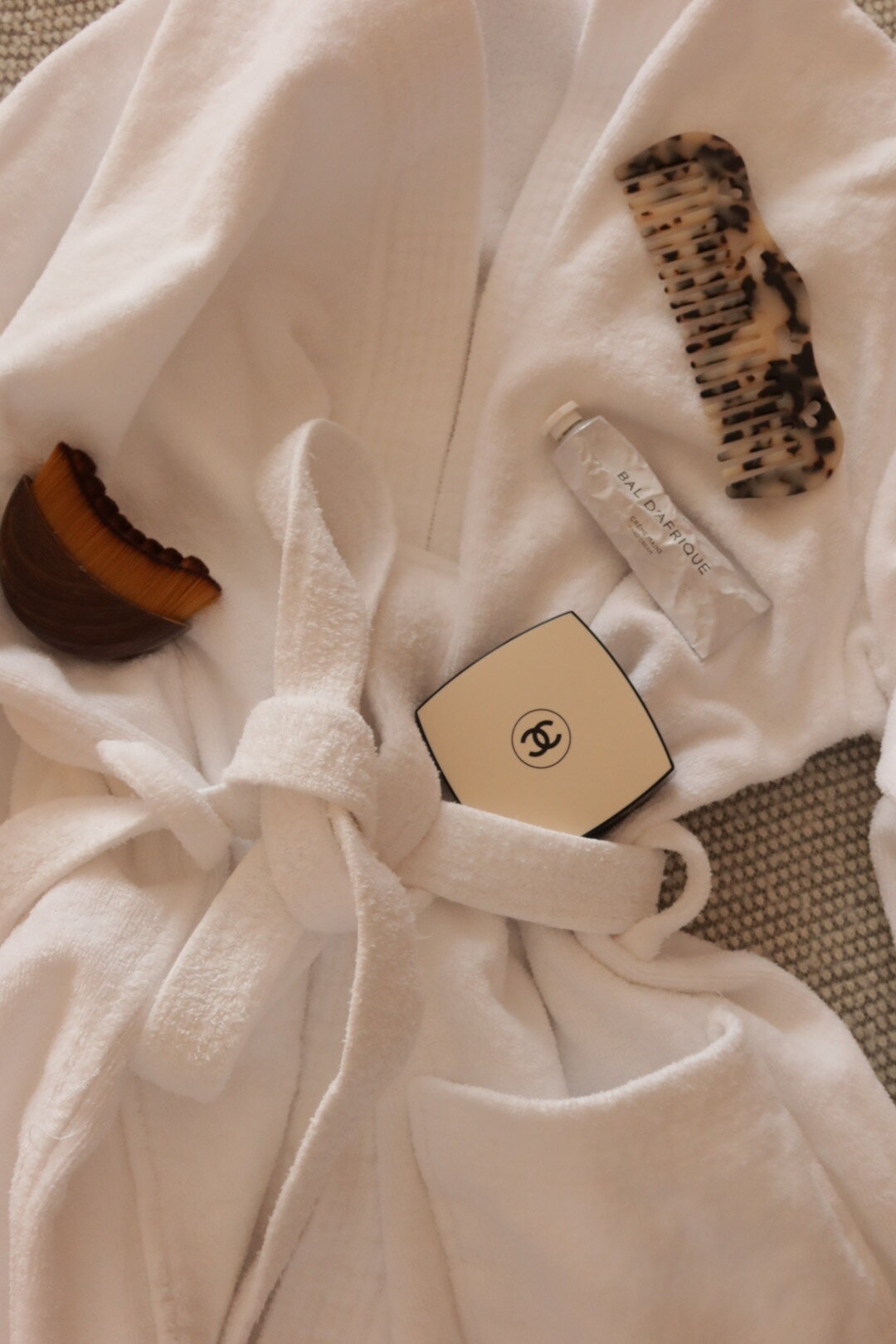 Flat lay of a white terry robe with a compact, hand cream, tortoiseshell comb, and makeup brush