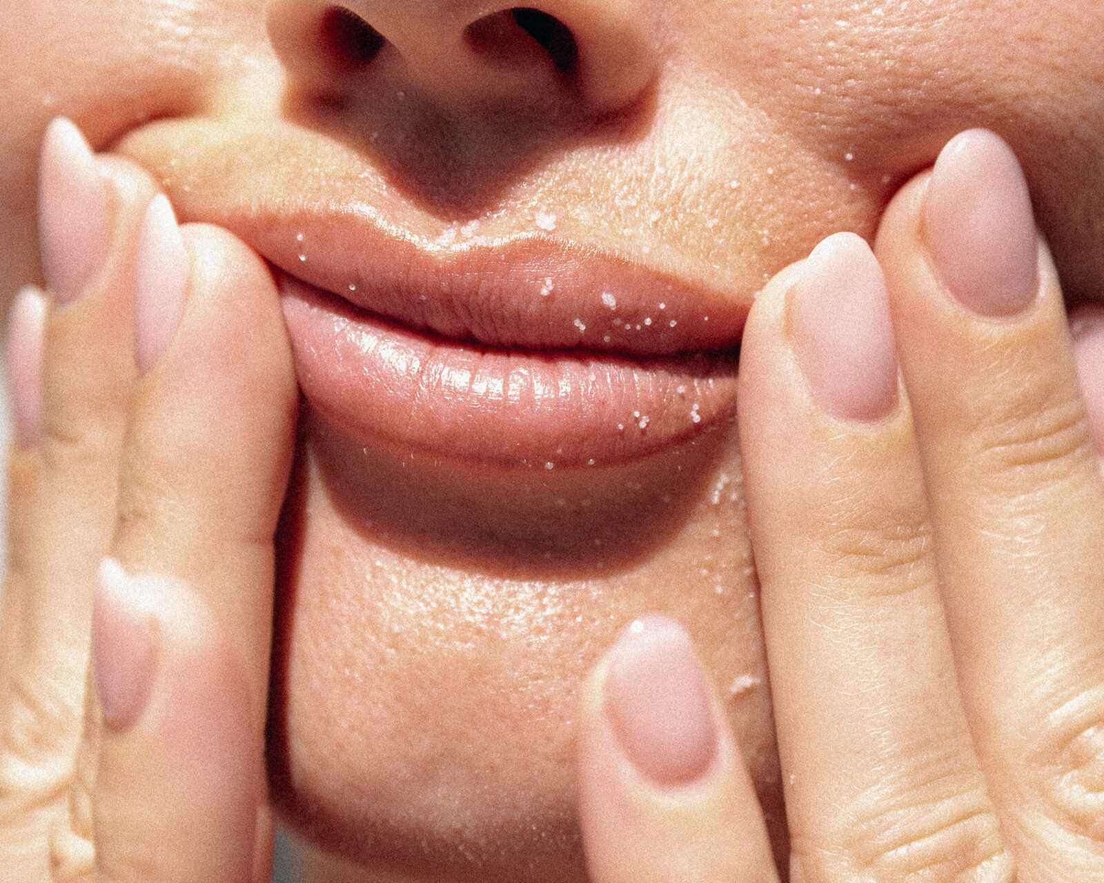 Close-up of full, glossy lips framed by fingertips with soft nude manicured nails