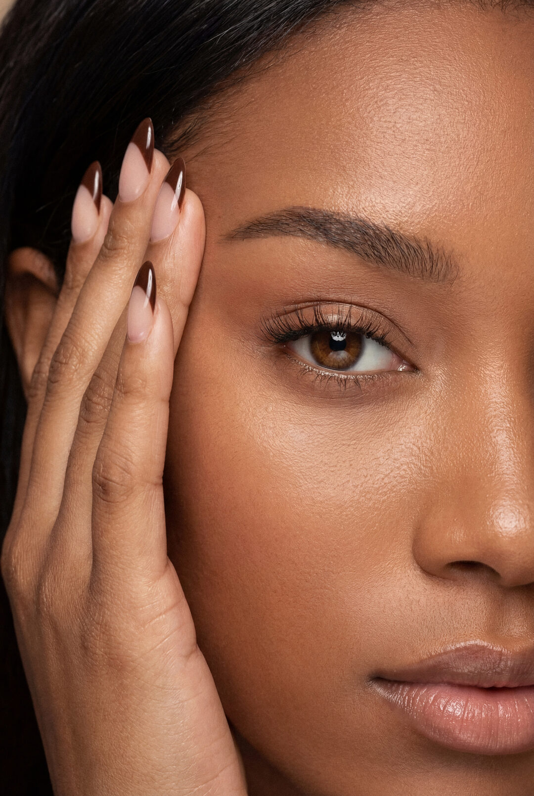 Close-up of a woman&rsquo;s eye and smooth under-eye area, fingertips with brown French-tip nails resting at her temple