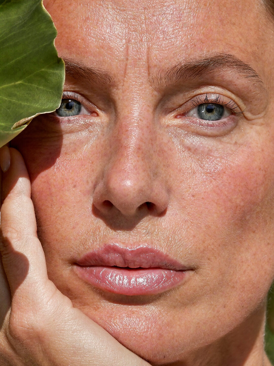 Close-up portrait of a woman with freckled, natural skin in sunlight, her hand resting on her cheek beside a green leaf