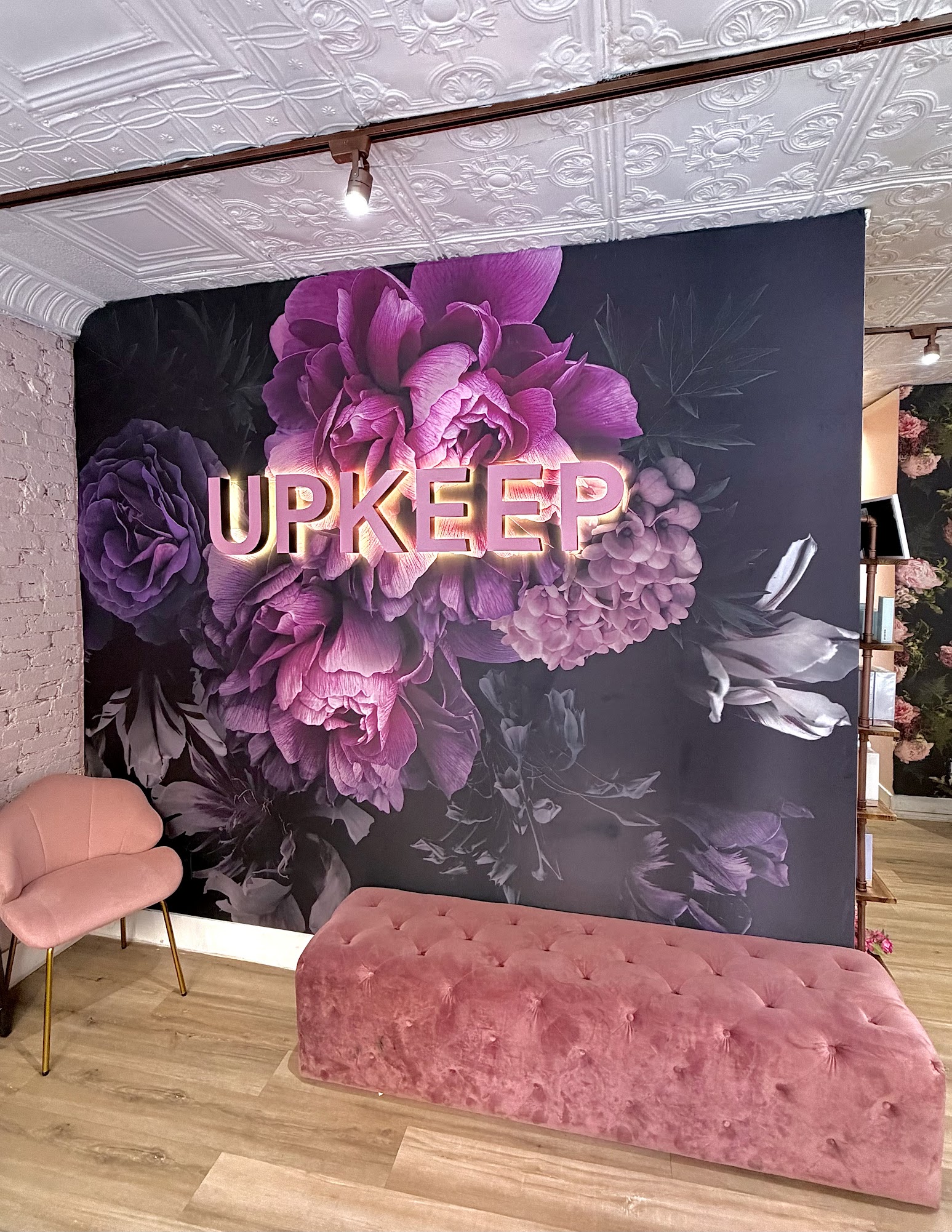Waiting area inside UPKEEP Med Spa Greenwich Village with a backlit UPKEEP sign on a purple floral mural above a pink velvet bench