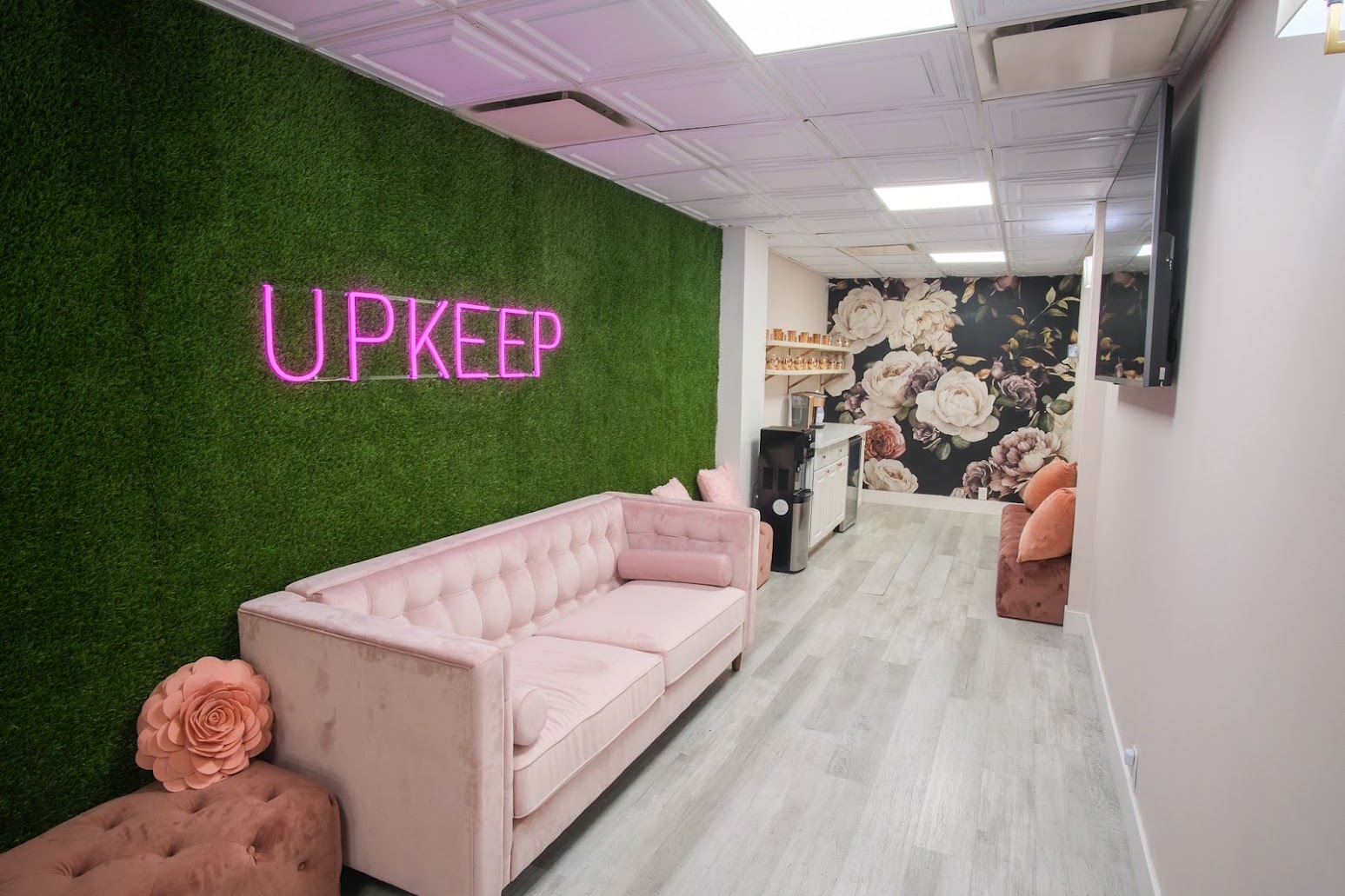 Lounge inside UPKEEP Med Spa Upper East Side with a pink neon UPKEEP sign on a green wall, a pink velvet sofa, and a floral mural